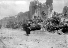 Une vieille femme passe devant l'église de Roncey en ruine, au pied de laquelle s'étend une file de blindés allemands détruits, 1er aout 1944